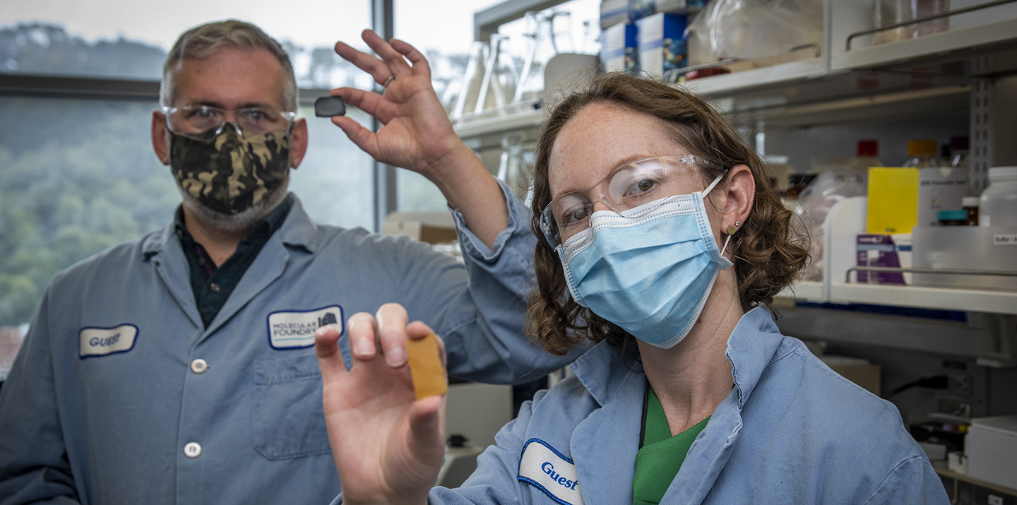 A man and woman in a lab wearing PPE holding small rectangular samples of PDK plastic